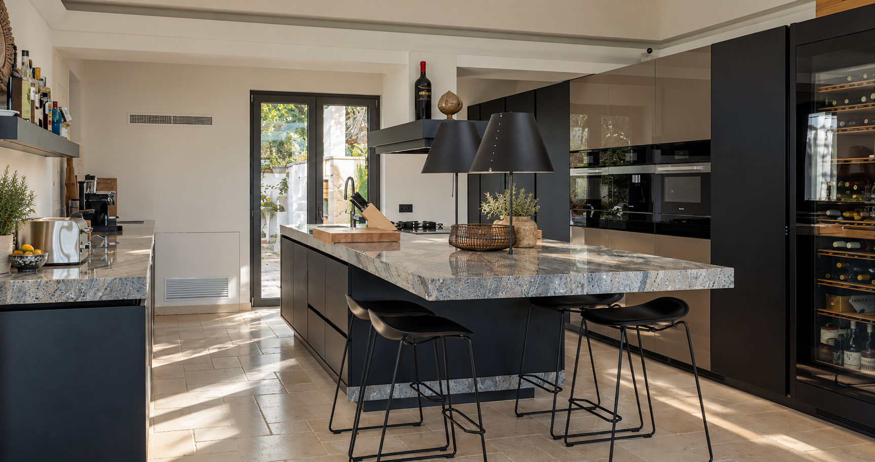 Modern kitchen with black cabinets, marble countertop island, bar stools, and a wine fridge, illuminated by natural light from glass doors.
