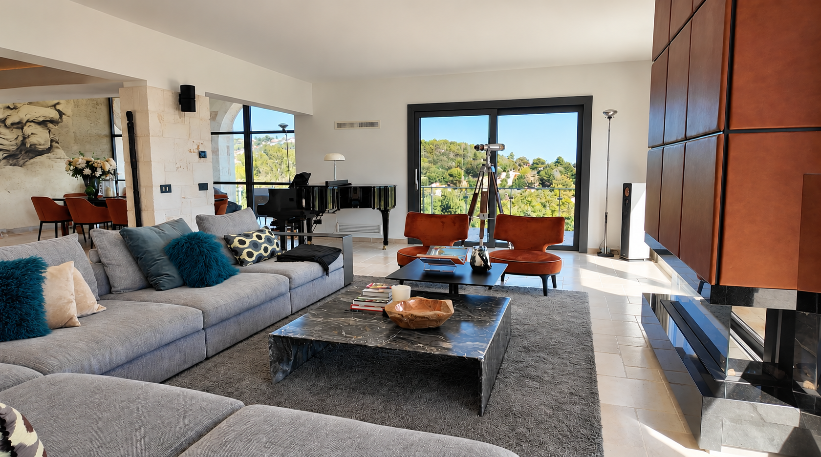 Living room with a gray sofa decorated with various colorful pillows, a black marble coffee table with a wooden bowl and candles, orange chairs, a large window with a view of green hills, a black grand piano, and a marble fireplace.