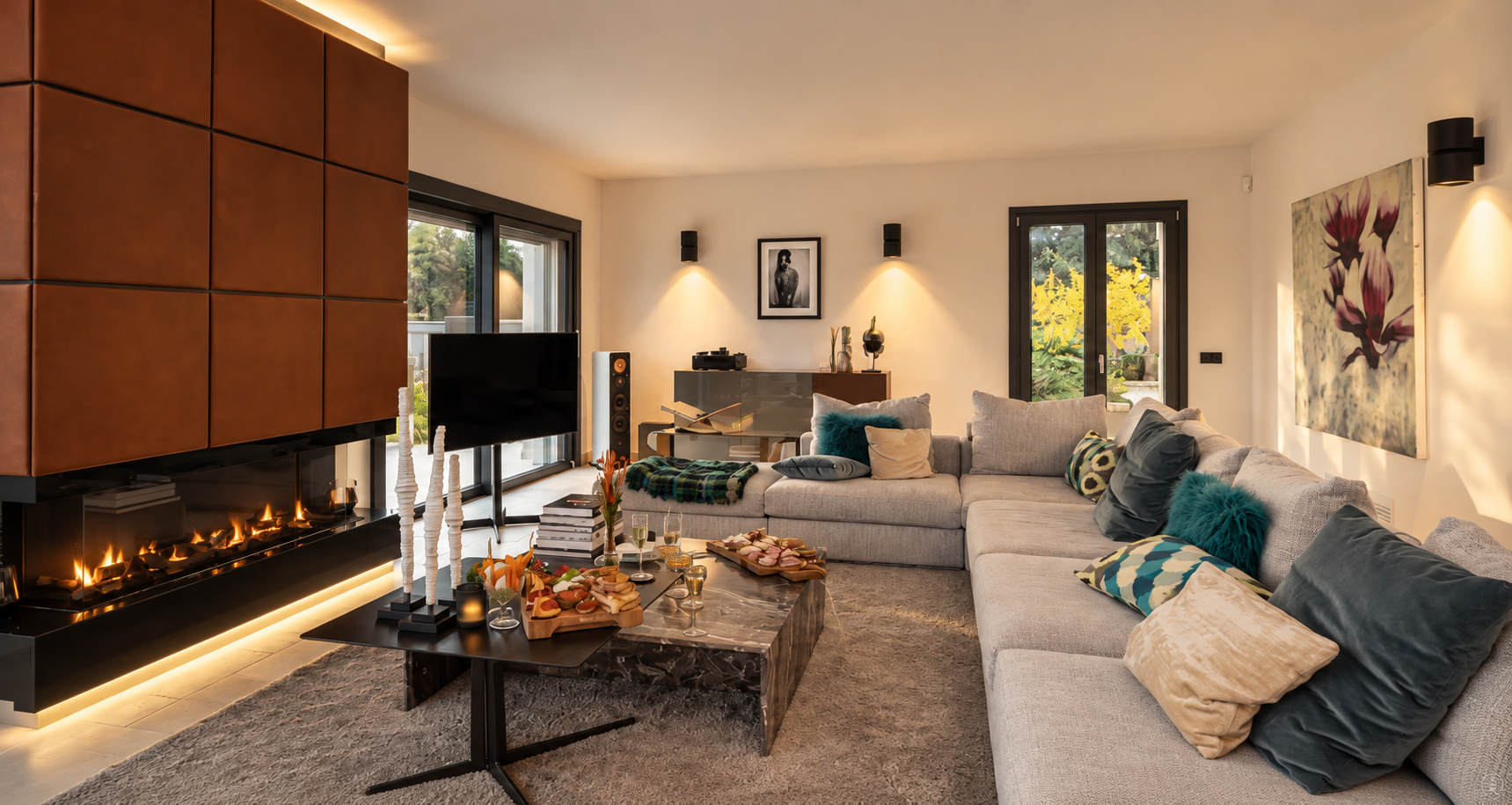 Living room with a beige sectional sofa with colorful cushions, a coffee table with snacks and drinks, a fireplace, a flat-screen TV, a large window and a sliding door, and modern artwork on the walls.