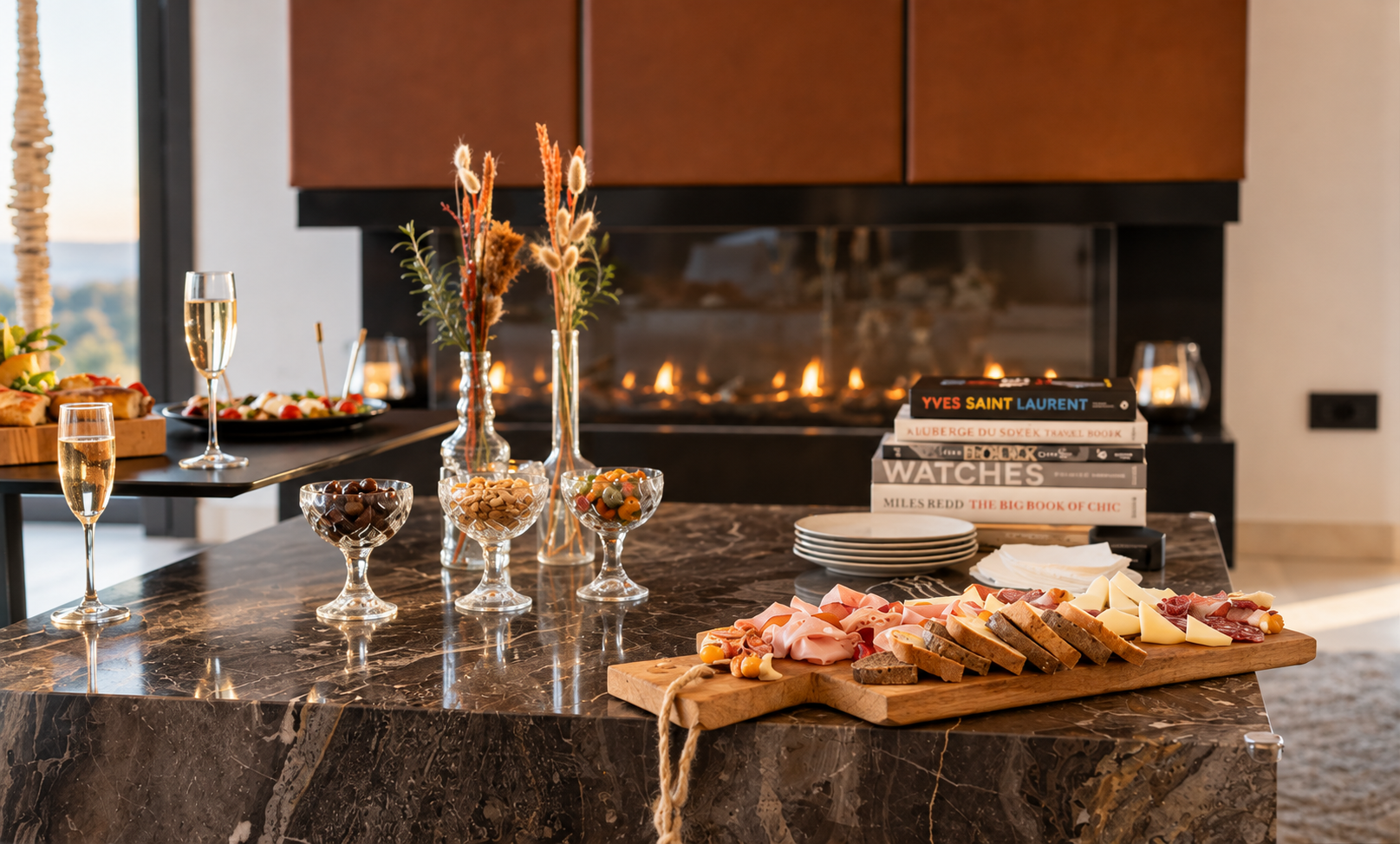 A marble kitchen island with a charcuterie board, bowls of candies, glasses of champagne, and stacked books, in front of a fireplace with flames and decorated with vases of dried flowers.