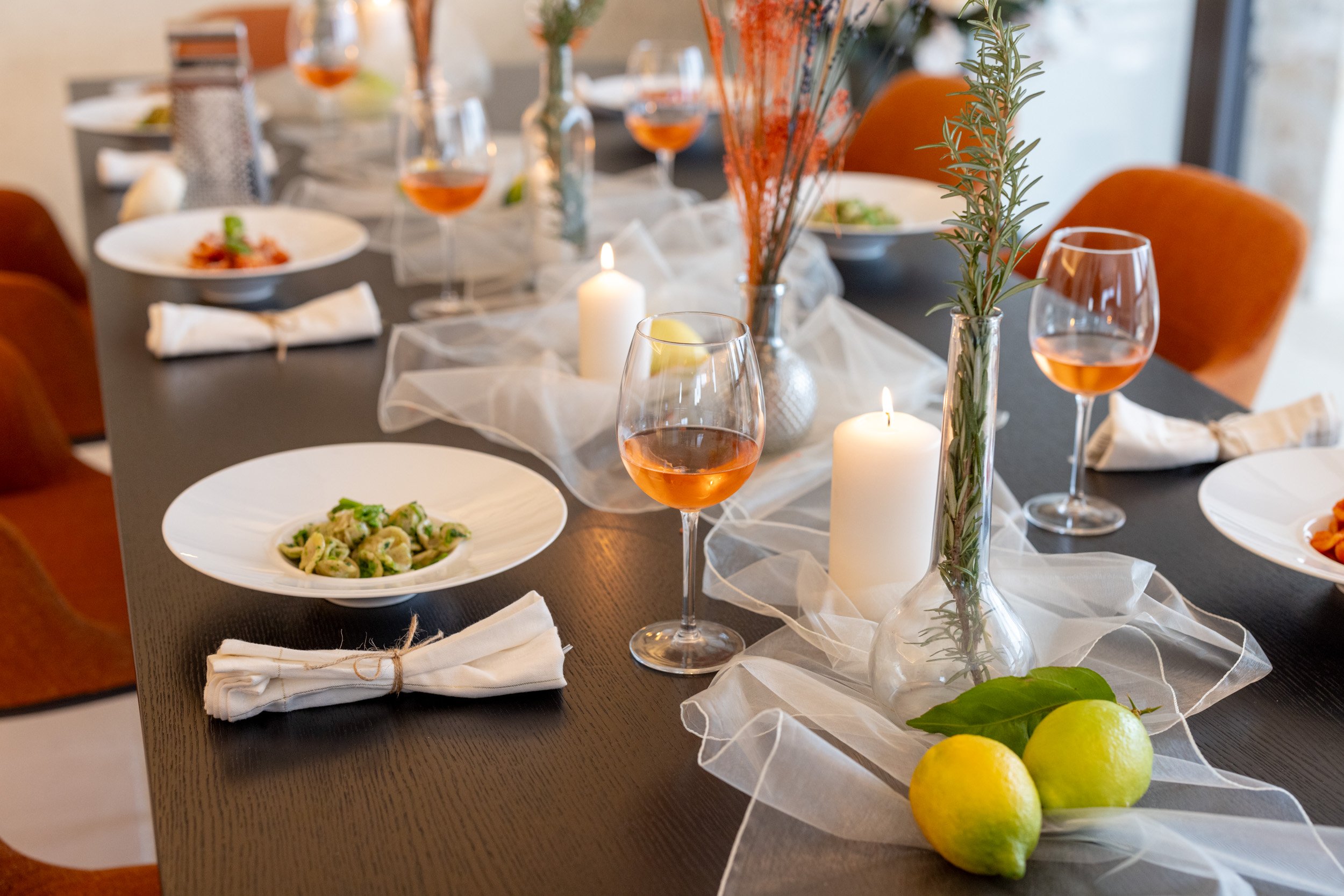 A decorated dinner table with plates of food, glasses of rosé wine, candles, and floral centerpieces, including lemons and green leaves.