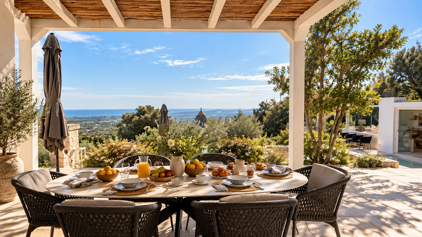 Outdoor patio with a round table set for breakfast, featuring bowls of fruit, a pitcher of orange juice, cups, and plates. The patio has a view of trees, greenery, and distant ocean, with clear blue sky.