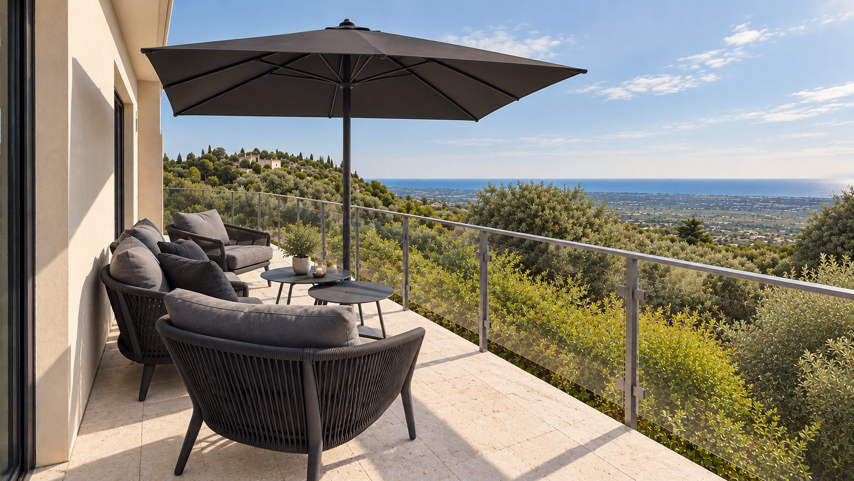 Outdoor balcony with black wicker furniture, potted plant, large black umbrella, railings, and a view of trees, hills, and ocean in the distance under a partly cloudy sky.