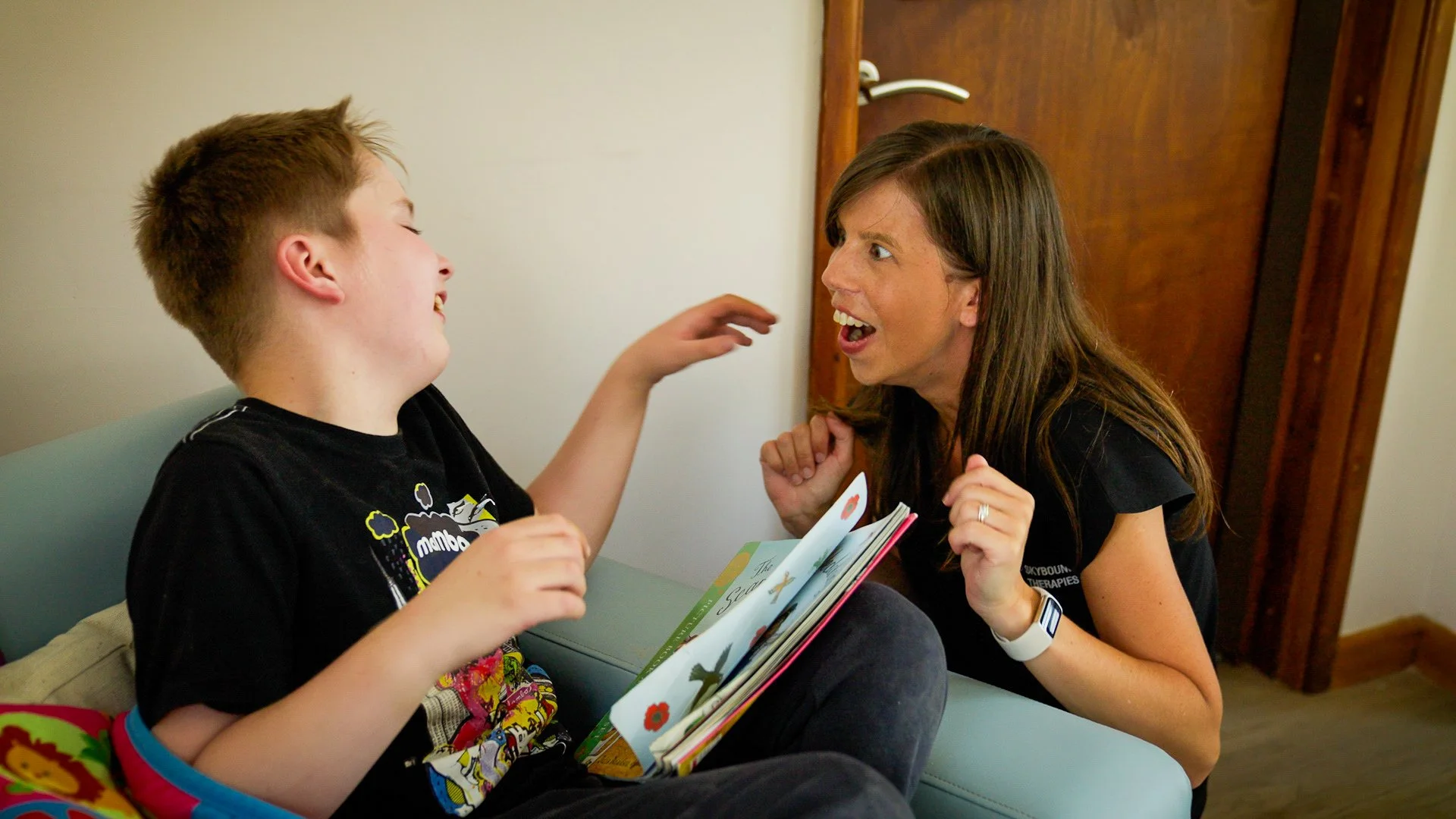 A young boy and a woman are sitting at a table, engaged in an animated conversation. The boy is holding a children's book, and both have expressive faces, indicating they are sharing a joyful moment.