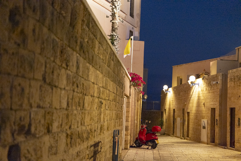 Nighttime view of a narrow alley with brick walls on either side, glowing wall-mounted lights, a red scooter parked in the alley, potted plants, and a yellow flag with pink flowers hanging from a window.