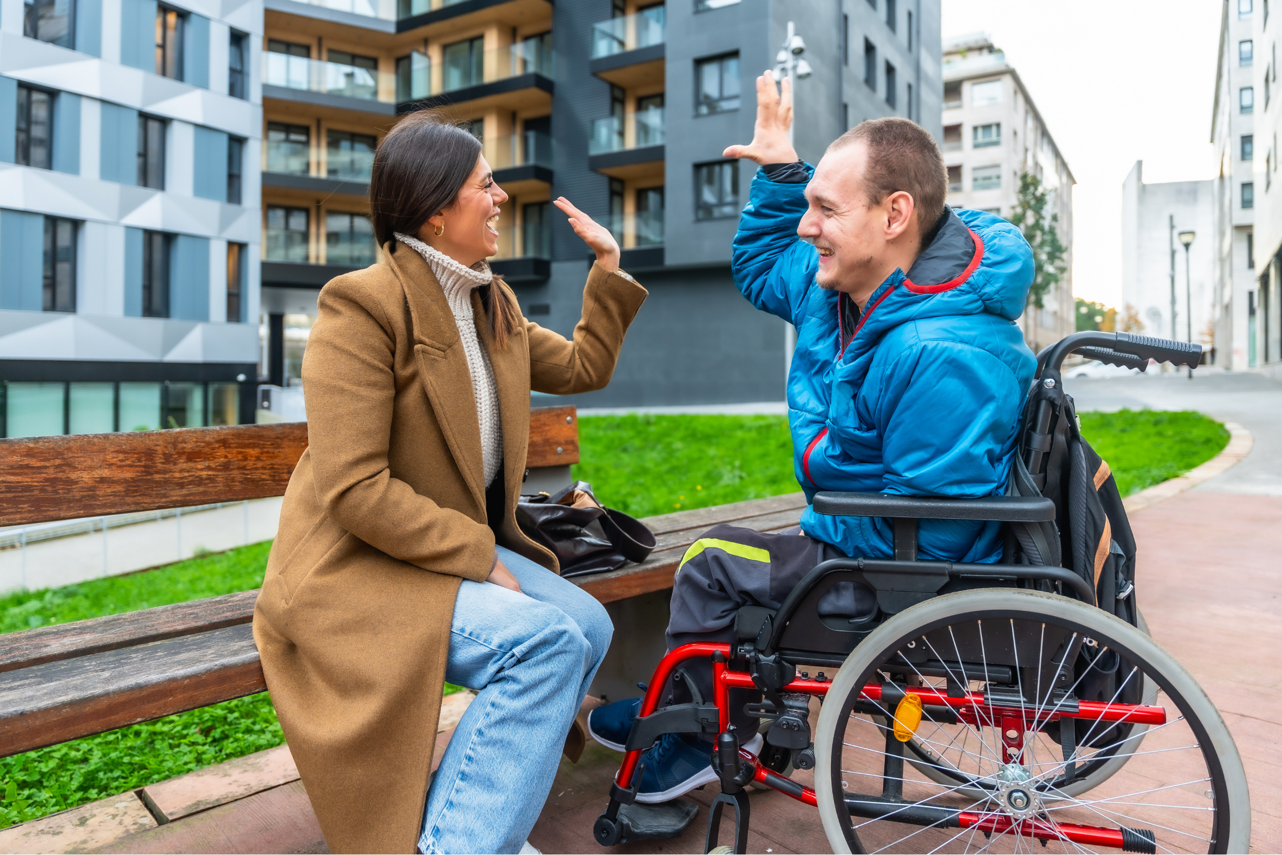 A woman and a man in a wheelchair sharing a joyful high-five while sitting on a park bench near modern apartment buildings.