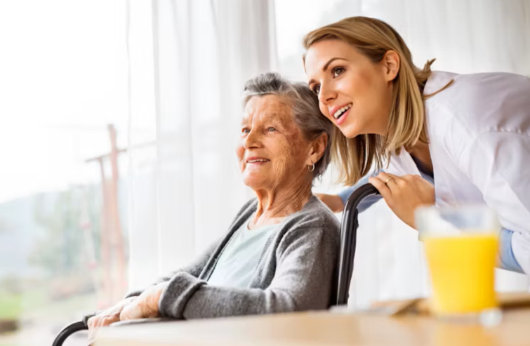A young female caregiver smiling and leaning over a wheelchair with an elderly woman sitting in it, inside a bright room.
