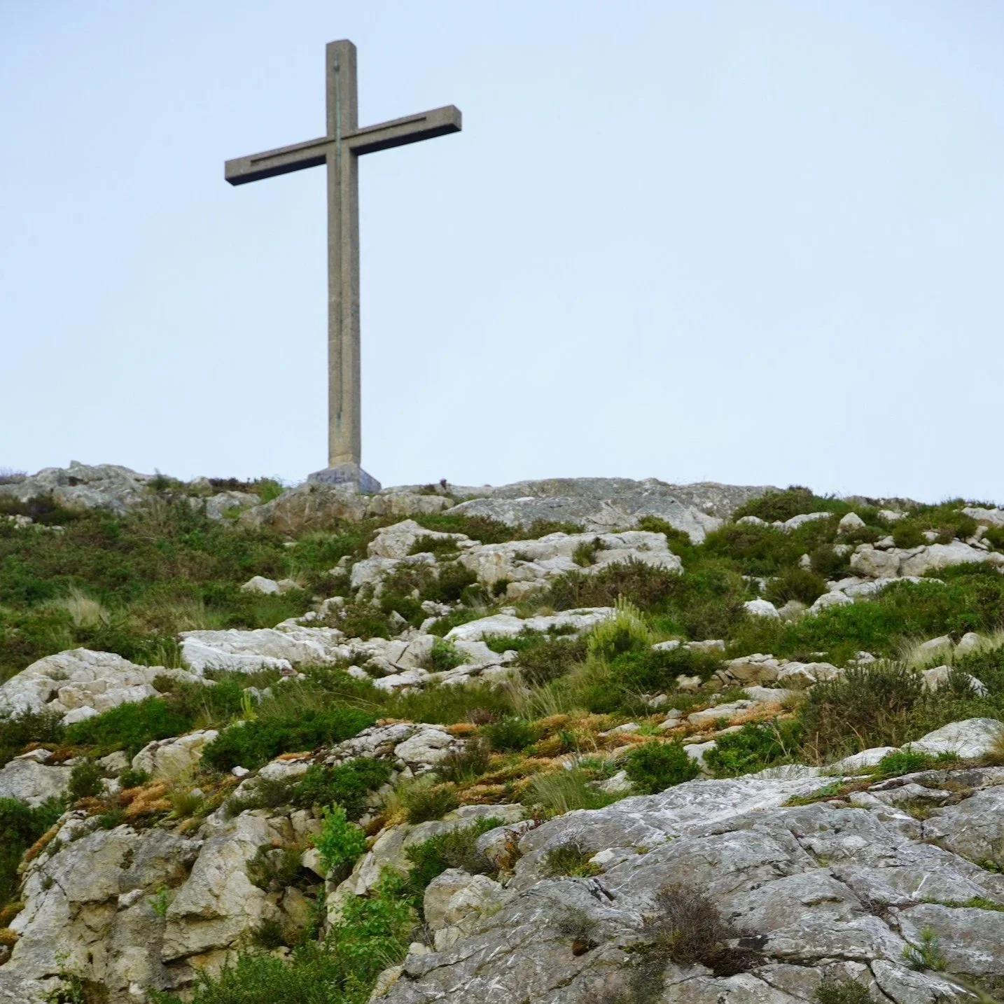 A large wooden cross standing on a rocky hilltop with green shrubs, overlooking a cloudy sky.