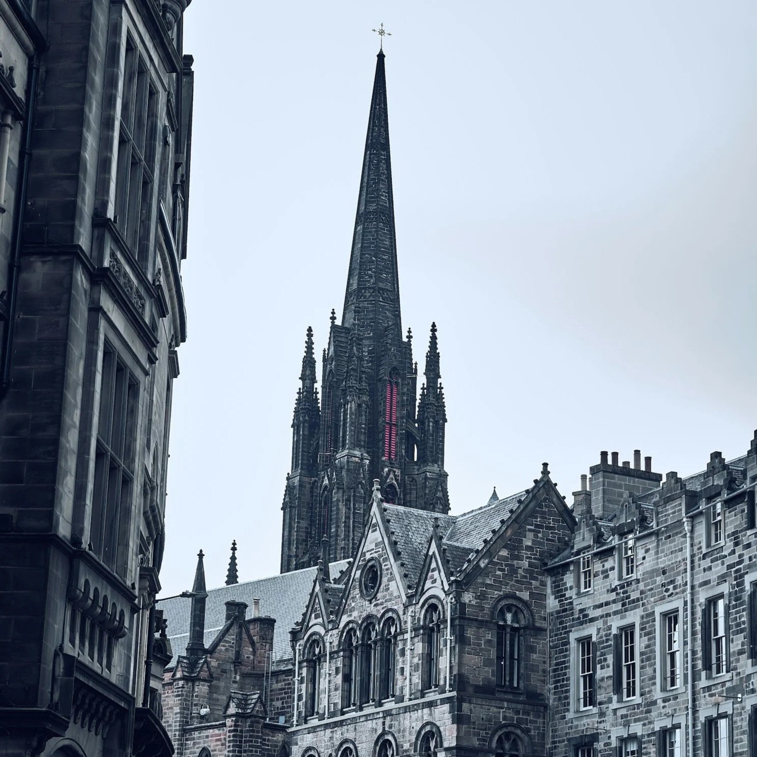 View of a city street with historic stone buildings and a tall gothic church spire with a cross on top.