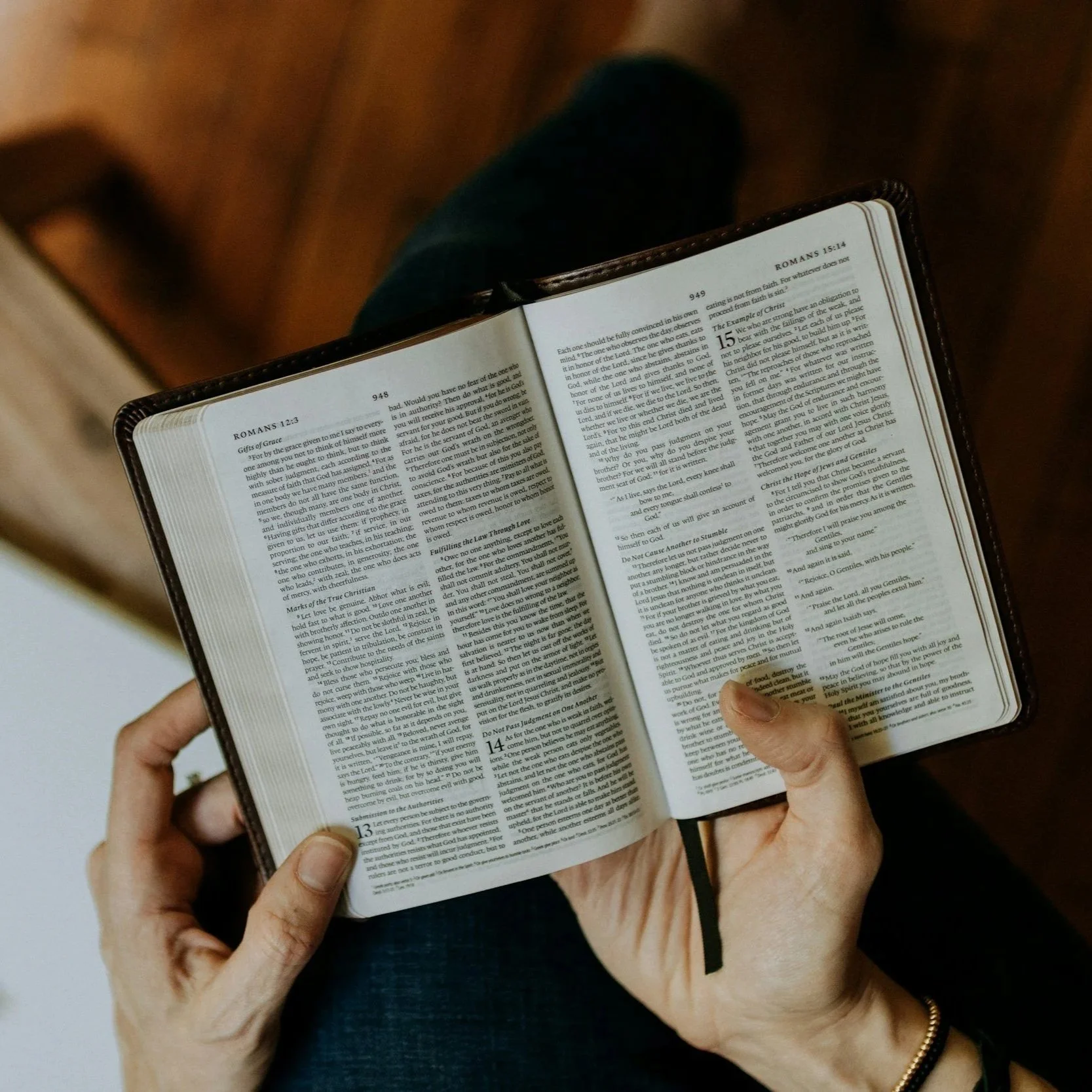 Person holding an open Bible, reading scriptures from Romans 12 and 15, with a wooden chair and a plain light-colored wall in the background.