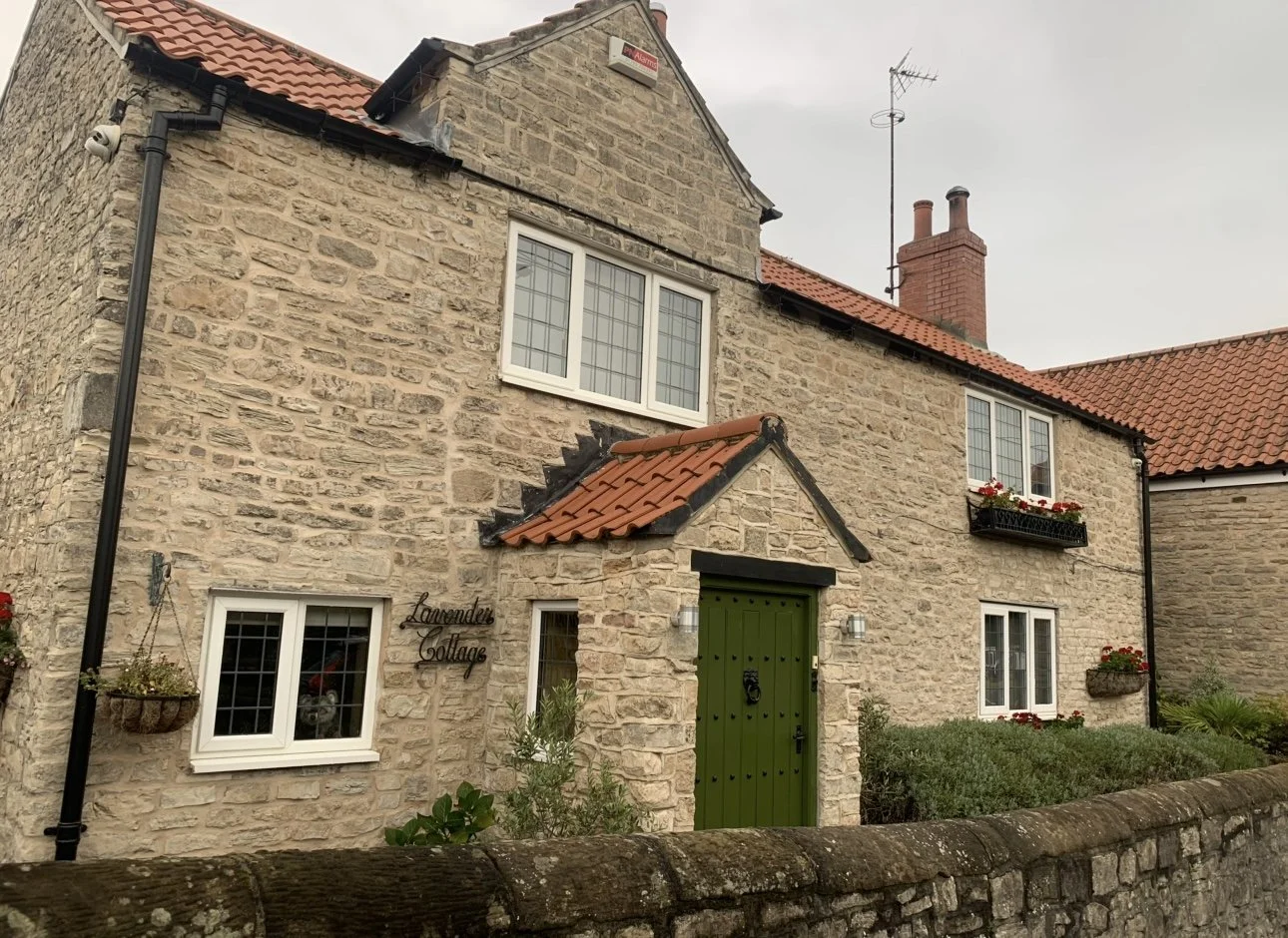 Stone cottage with a green door, white-framed windows, flower planters, and a sheared hedge in front.