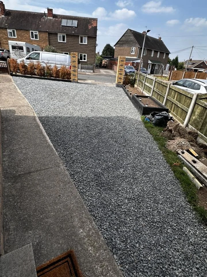 A gravel driveway under construction in front of a residential house, with a brick entrance, a fence on the right, and neighboring houses and parked cars visible in the background.