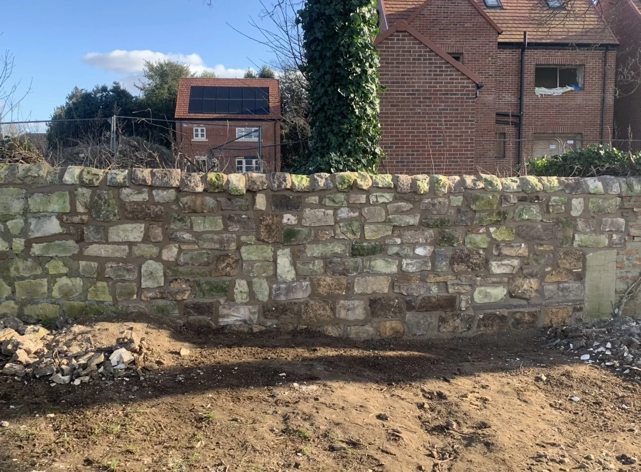 A stone wall with a dirt ground in front, behind it is a house with brick walls and a red roof, and another house with solar panels on its roof in the background. The sky is blue with a few clouds.