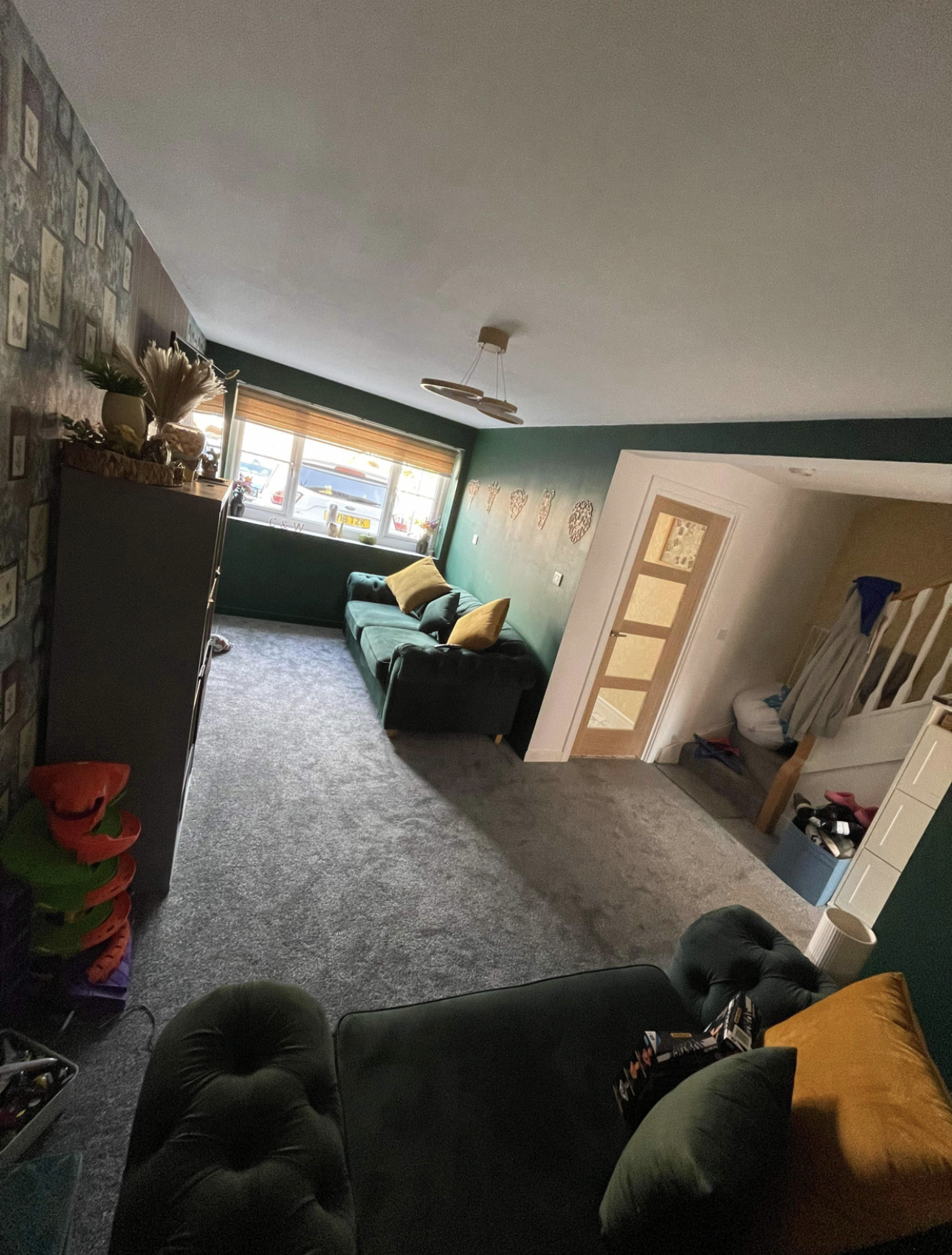 A cozy living room with a gray carpet, dark green sofa with yellow pillows, and a window with wooden blinds letting in natural light. The wall has decorative hearts, and there's a staircase with a white railing nearby. A black armchair with a yellow pillow is in the foreground, and a tall white storage unit is on the right.