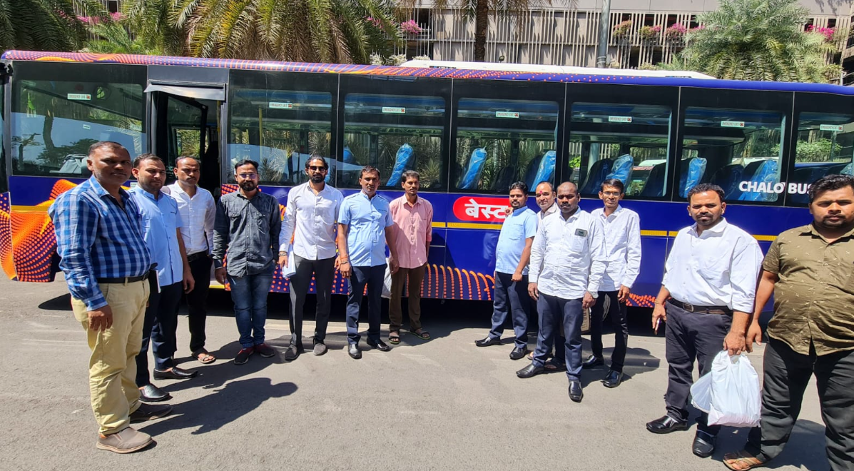 A group of men standing in front of a blue bus with Hindi writing, outdoors with palm trees and buildings in the background.