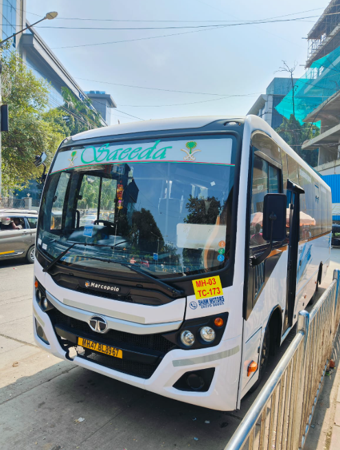 White bus with the name 'Sareeda' and a palm tree logo on the front windshield, parked on a street with other vehicles and buildings in the background. Apex Commute