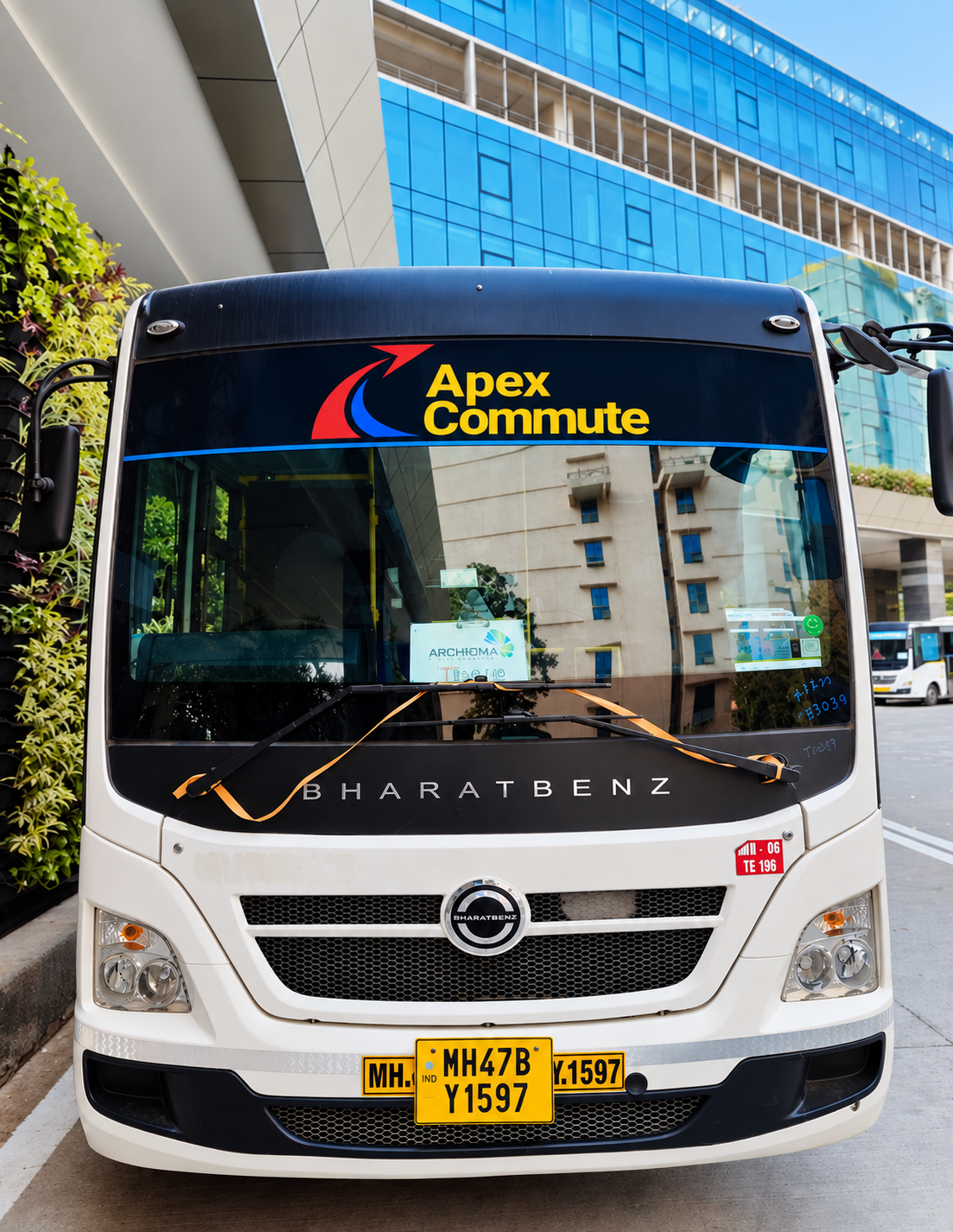 A white Bharat Benz bus with a black banner that says 'Apex Commute' parked in front of modern glass buildings in an urban area.