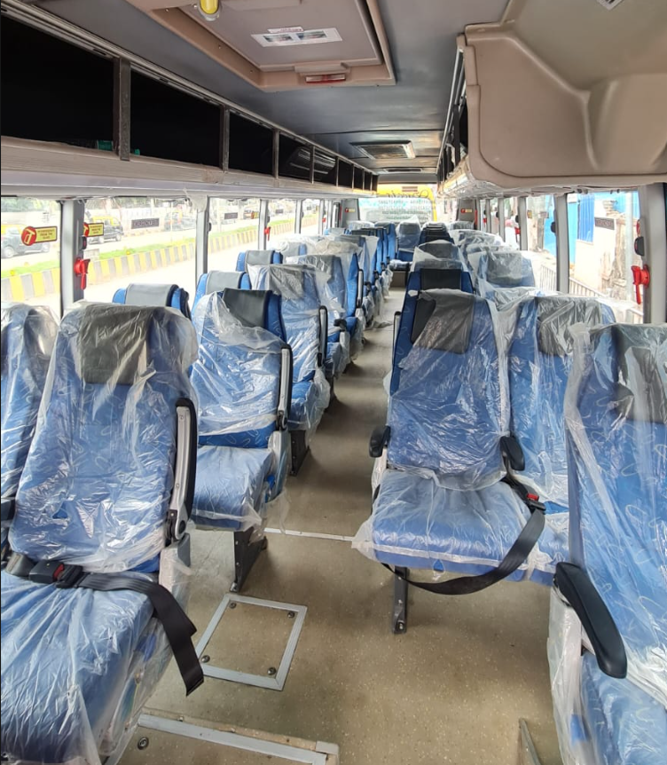 Inside of a bus with blue seats covered in plastic, parked near a street with a yellow and black barrier, and buildings visible outside.