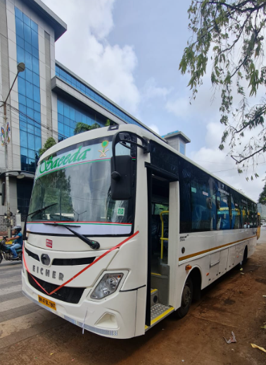 A white bus with the name 'Encerada' written on the front, parked outdoors in front of a modern glass building, with a tree on the right and a partly cloudy sky.