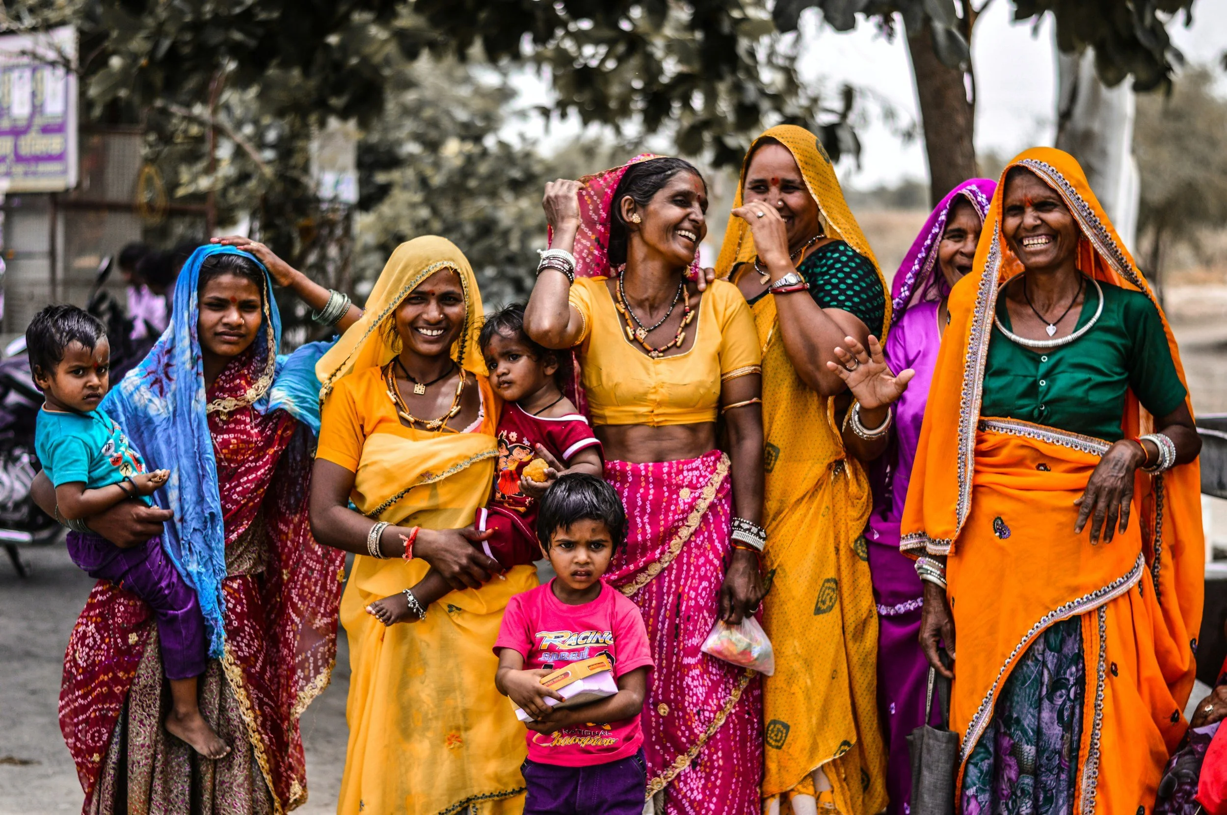 Group of women and children dressed in colorful traditional Indian clothing, smiling and gathered outdoors under trees.