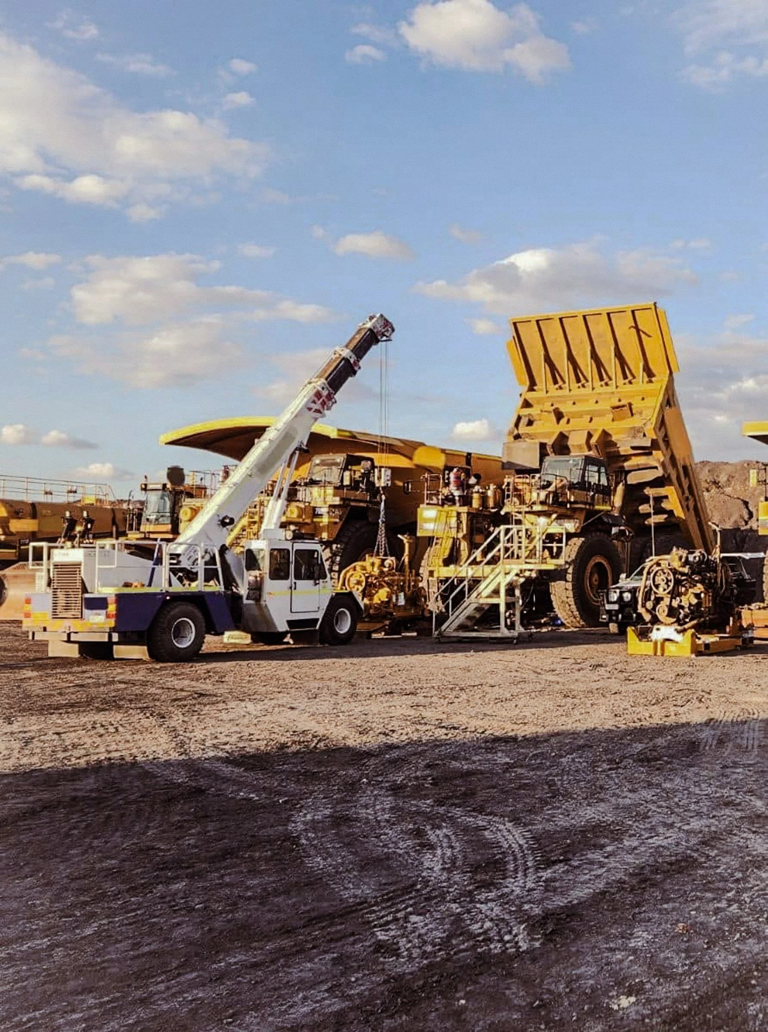A large yellow mining dump truck being loaded by a white crane at a mining site with a cloudy sky in the background.