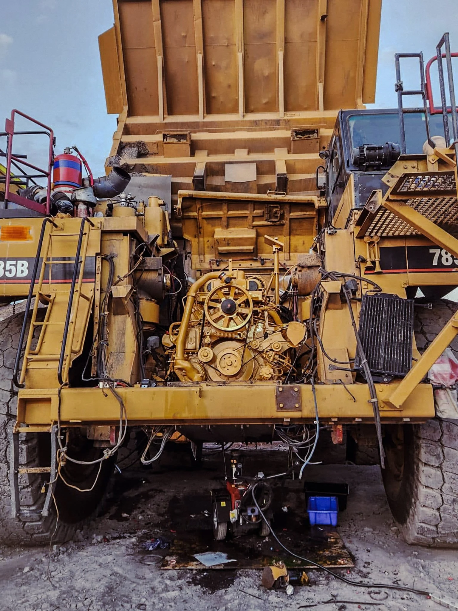 A large yellow construction machine with wires and parts exposed, situated outdoors on a dirt ground, with a stone wall and cloudy sky in the background.