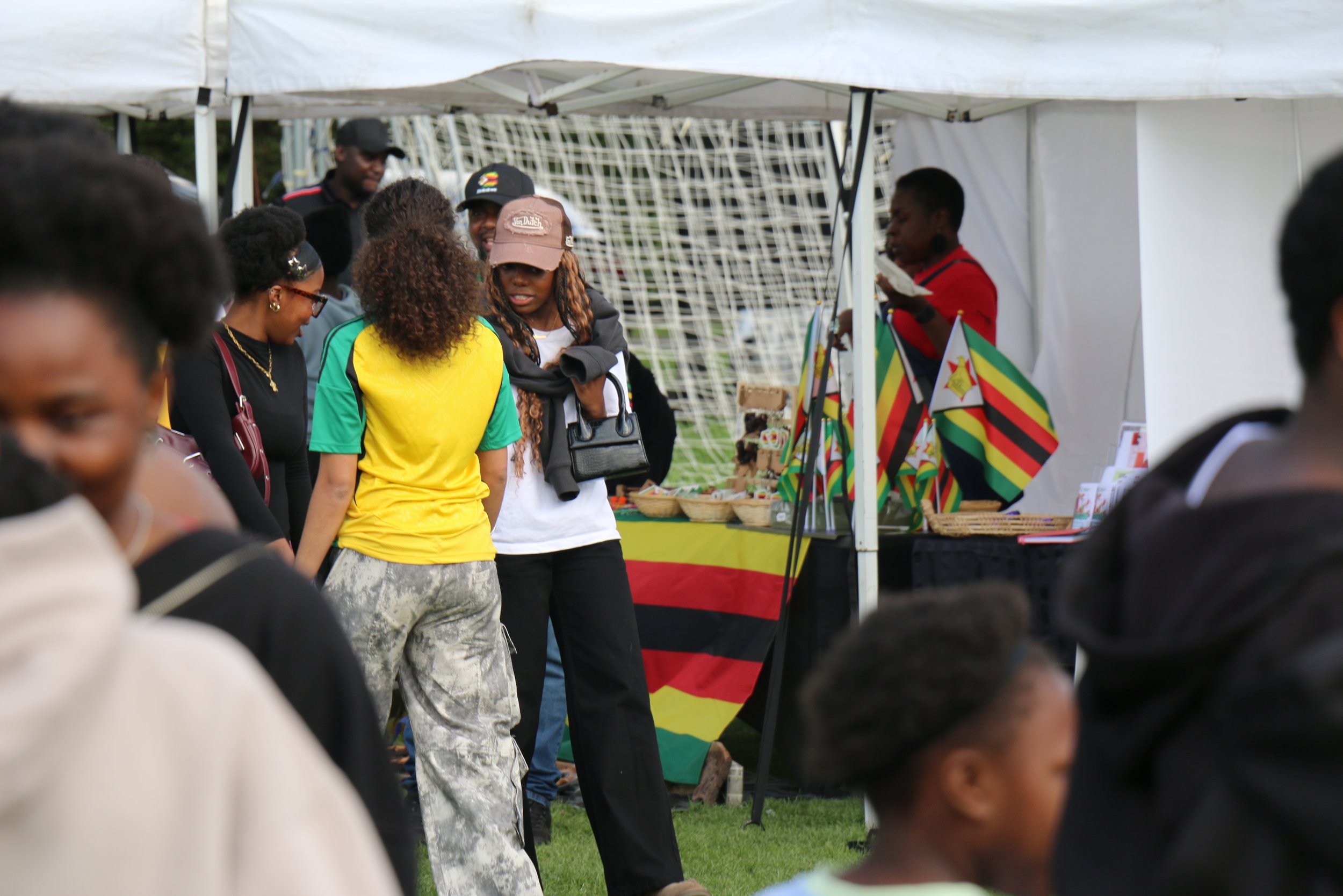 People gathered around a booth at an outdoor event, with some individuals looking at handmade crafts and flags representing Zimbabwe.
