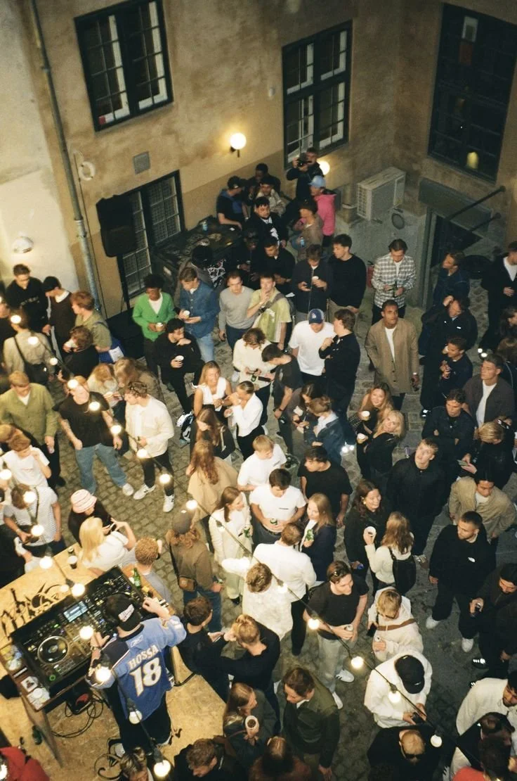 People gathered at an indoor party with string lights, some holding drinks, and a DJ playing music.