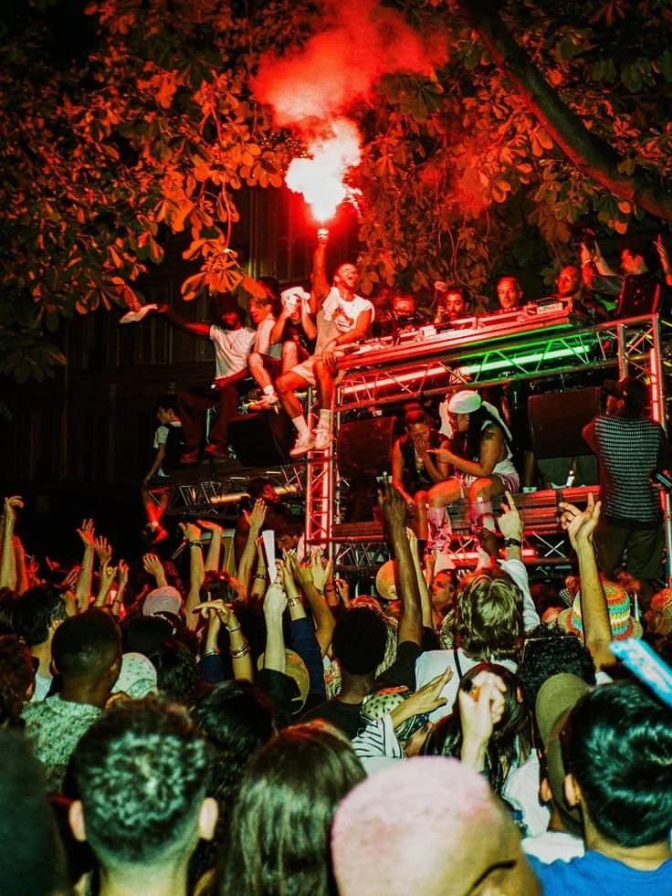 Crowd enjoying a lively outdoor music event at night, with performers on a platform and one person holding a flare emitting bright light.