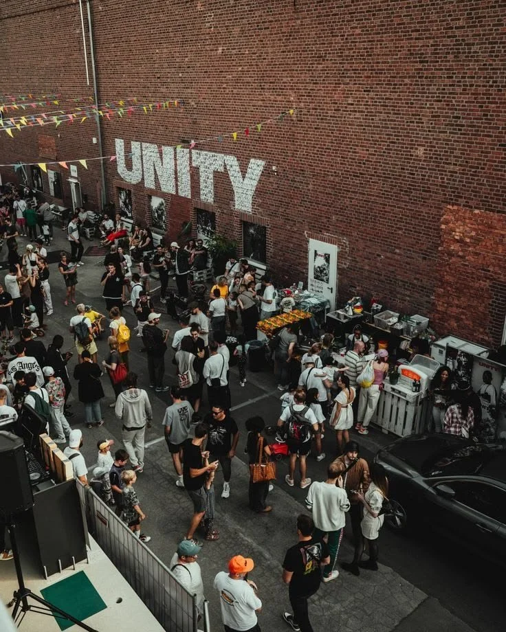 People gathered outside of food trucks and booths in an alleyway, with a large brick wall in the background featuring the word