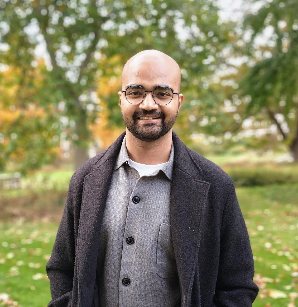A smiling man with glasses and a beard wearing a gray shirt and black coat standing outdoors in front of trees with autumn leaves.
