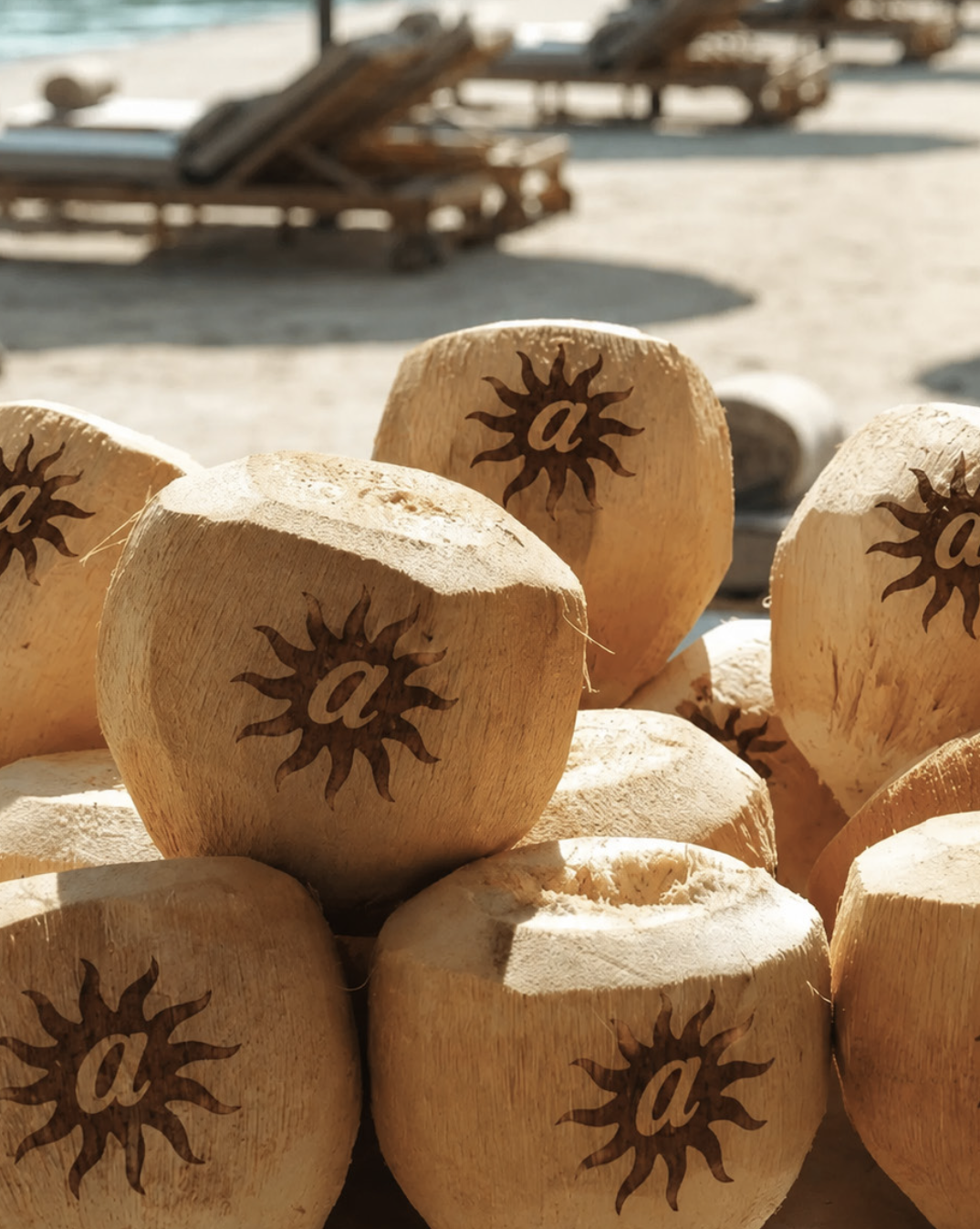 Several wooden coconuts with a logo of a sun and the letter 'a' burned or painted on them, stacked on a beach with lounge chairs and sandy ground in the background.