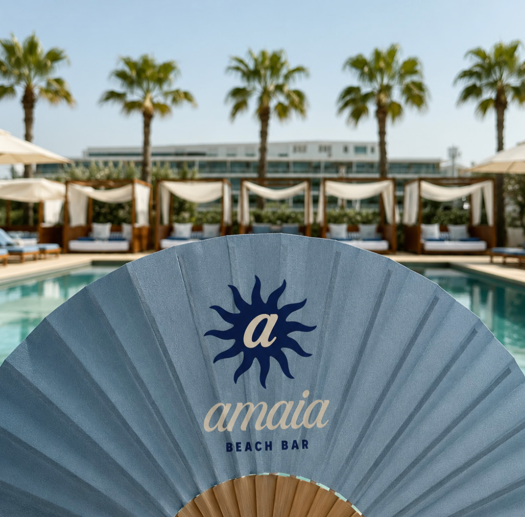 A blue paddle fan with the words 'a Maia Beach Bar' and a sun logo, held in front of a poolside scene with cabanas, lounge chairs, umbrellas, palm trees, and a modern building in the background.