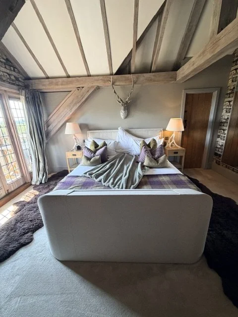 Cozy bedroom with a platform bed, decorative pillows, and a sloped ceiling with exposed wooden beams. Large window with curtains and nightstands with lamps on either side.