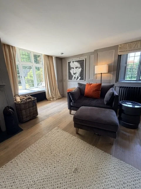 Living room with large windows, curtain drapes, a gray armchair with a matching ottoman, an orange pillow, a framed black-and-white portrait on the wall, a floor lamp, a black side table, and a beige rug.