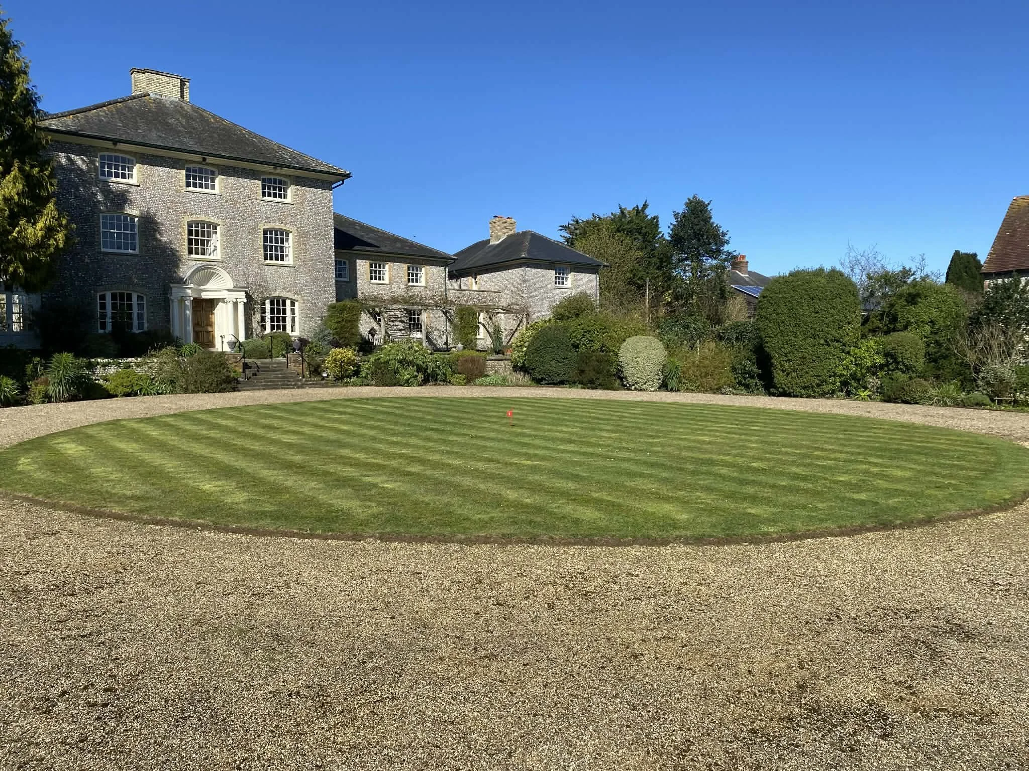View of a large, historic brick house with many windows, surrounded by well-maintained garden and trees, under a clear blue sky.