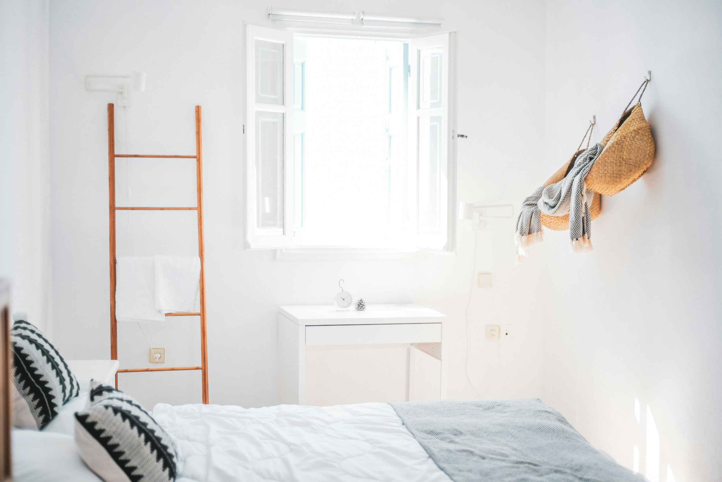 A bright bedroom with a bed in the foreground, black and white patterned pillows, an open window letting in sunlight, a wooden ladder-style towel rack with a white towel, a small white table with decorative items, and a wall with hanging woven bags and scarves.