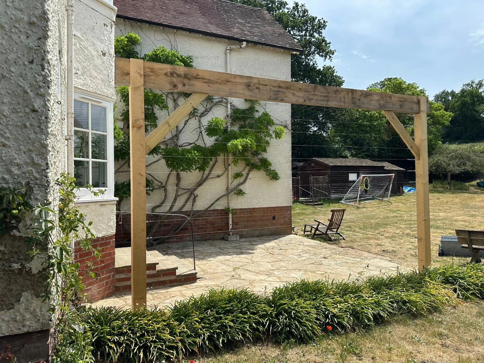 A backyard with a newly built wooden structure for a patio or pergola, adjacent to a house with a window and white stucco walls. There is a stone-paved patio area, a lawn with some benches and a children's soccer goal, and a small wooden shed in the 