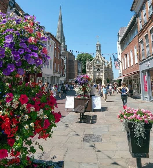A busy pedestrian street lined with colorful hanging flower baskets, with an ornate historic building and a tall spire in the background, under a clear blue sky.