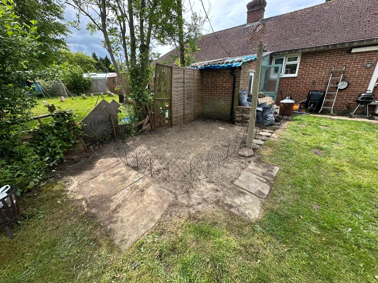 Backyard with dirt area enclosed by small metal fence, adjacent to paved patio, with some tools, bags, and outdoor items near the brick house, and a grassy lawn with trees and a clothesline in the background.
