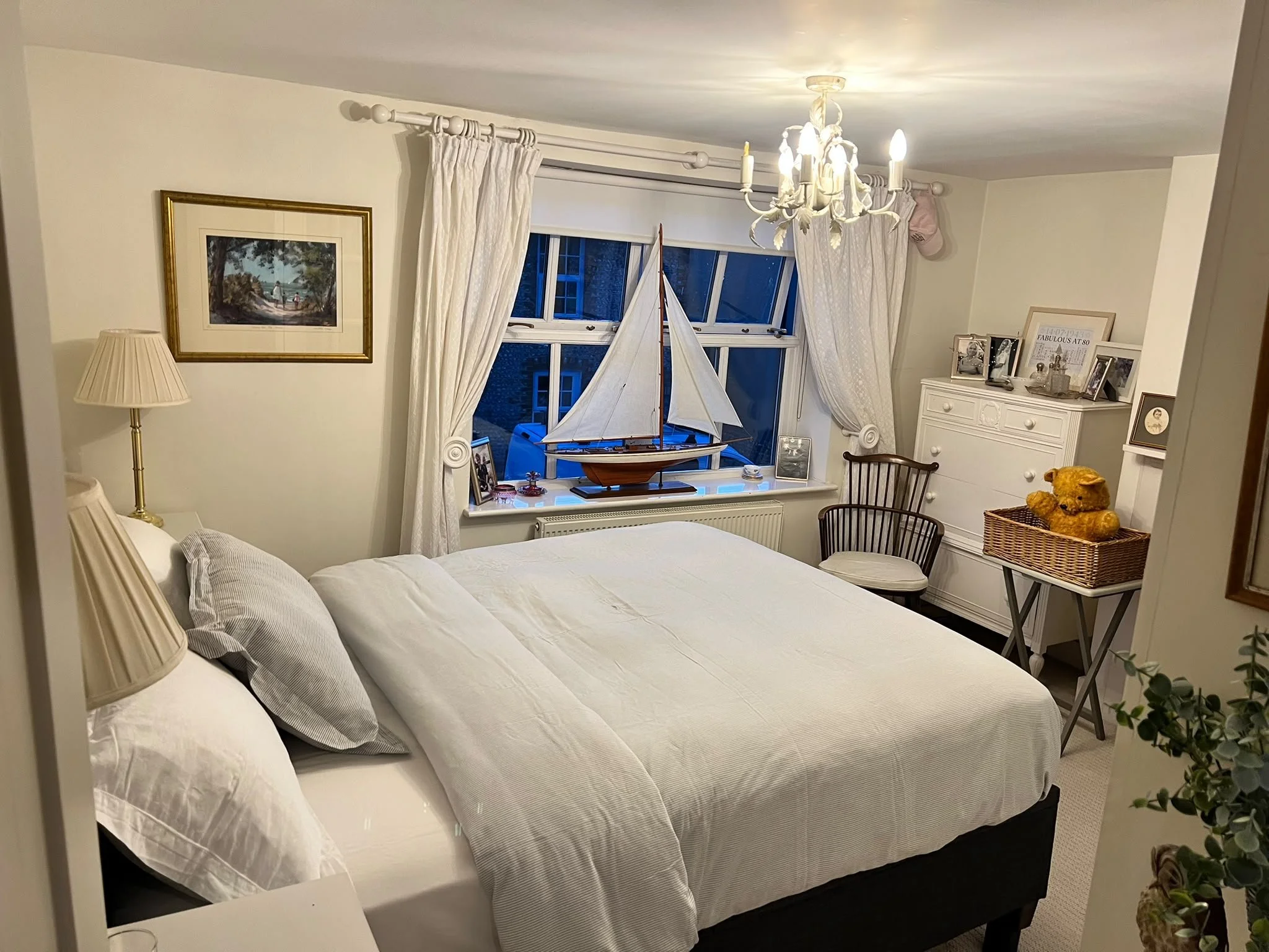 A cozy bedroom with a white bed, beige lamps, framed pictures, and a model sailboat on the windowsill, with cream-colored curtains and a chandelier overhead.