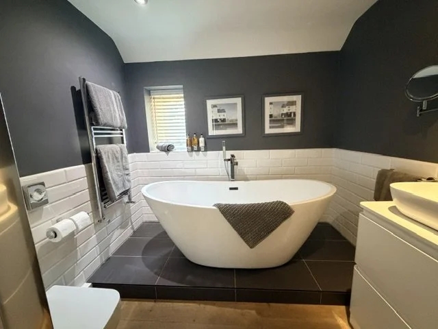 Modern bathroom with a white freestanding bathtub on black tiled floor, dark gray walls, and white subway tile halfway up; small window with blinds, two framed pictures above; towels hanging on a wall-mounted heated towel rack; vanity with sink on th