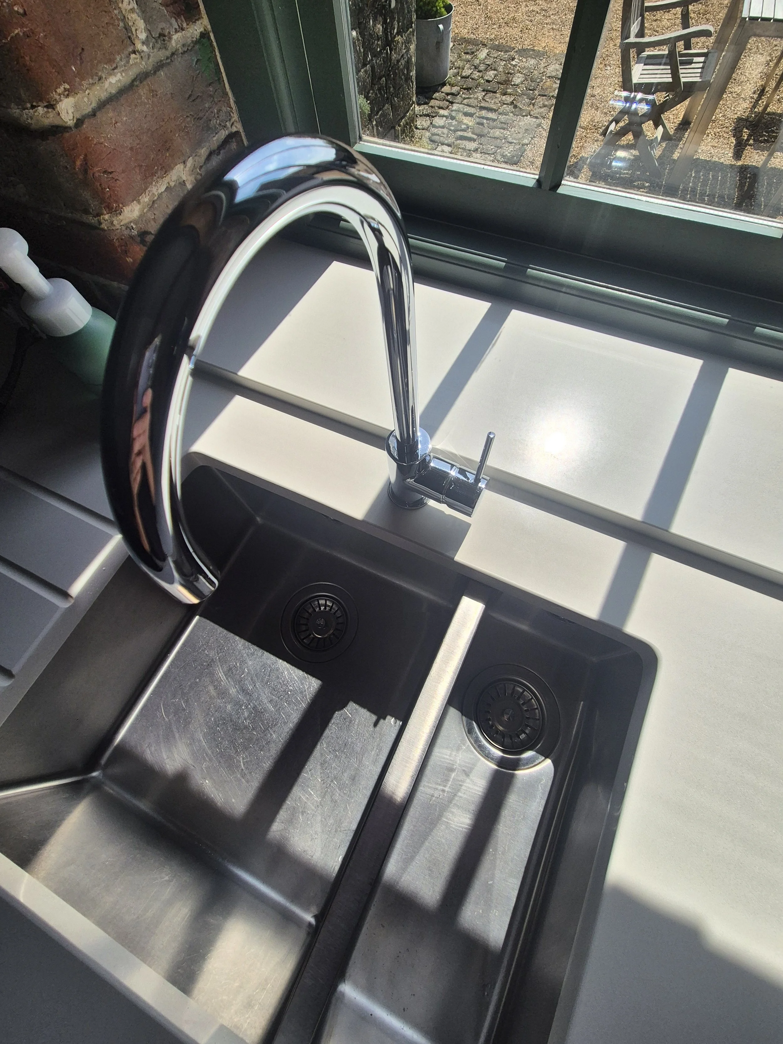 Indoor stainless steel kitchen sink with a faucet and a drain, sunlight casting shadows on the countertop, window with a view of an outdoor patio with cobblestone ground and a wooden chair.