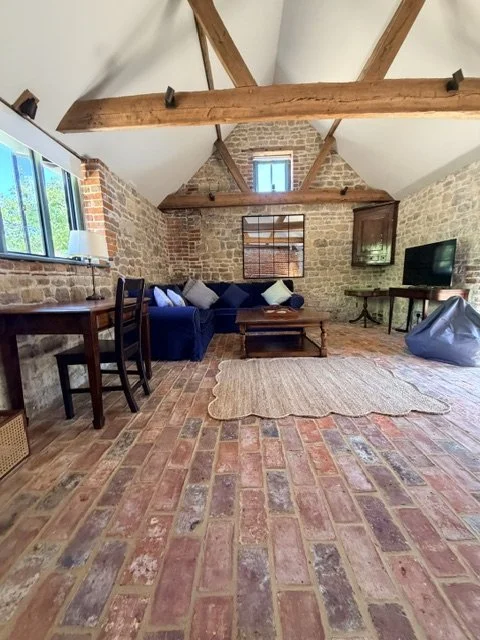 A rustic living room with brick flooring, stone walls, and exposed wooden beams. There is a blue sofa with pillows, a wooden coffee table, a side table, a bean bag chair, and a television. Large windows allow natural light into the space.