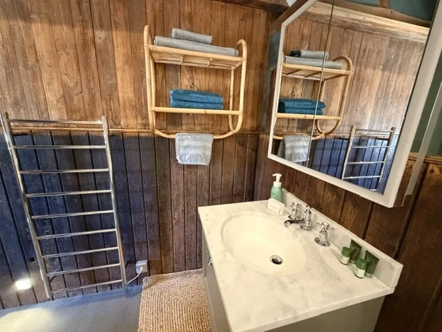 Bathroom with wooden wall paneling, a white marble sink vanity, a mirror, a soap dispenser, and a towel shelf with folded towels. Towel racks are on the wall and floor.