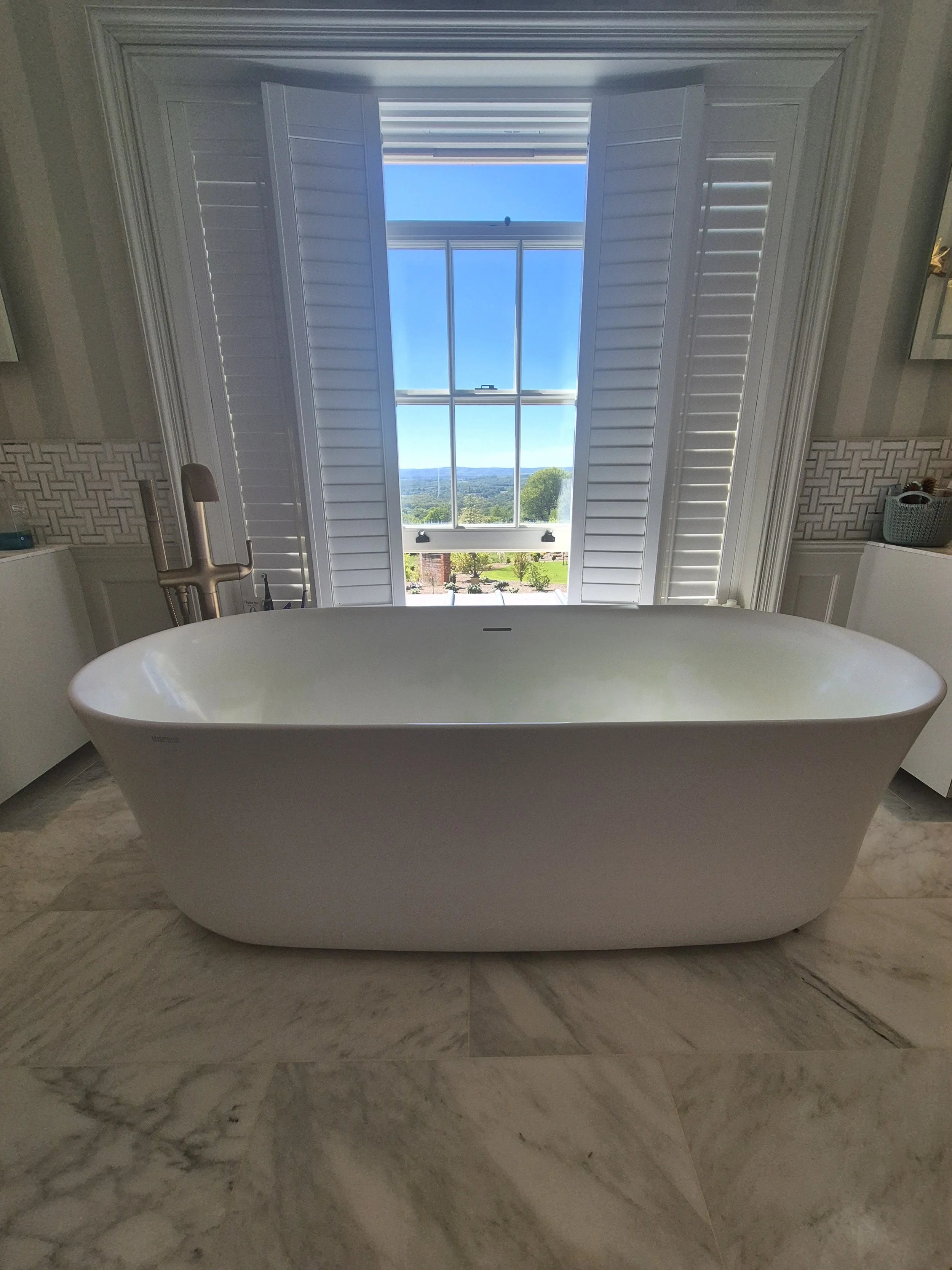 A white freestanding bathtub in a bathroom, positioned in front of a large window with open white shutters showing a scenic outdoor view of trees, hills, and a blue sky.