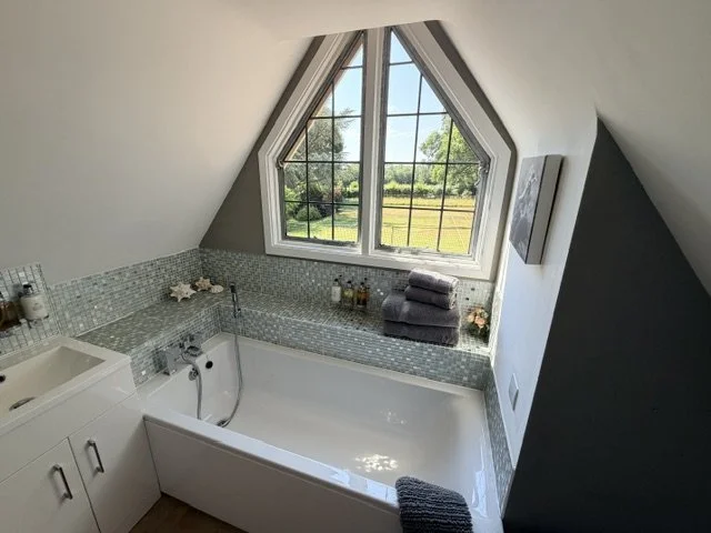 Bathroom with a window overlooking a green outdoor landscape, built-in tub with a gray towel draped over the edge, small cabinet with toiletries, folded towels, and a flower on the ledge, and gray and white mosaic tile surround.