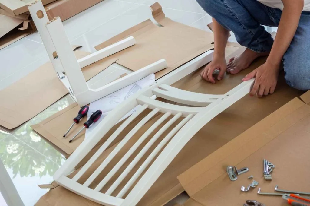 A person assembling a white wooden chair or stool with slatted seat, on a brown paper-covered floor, with tools like screwdrivers and screws nearby.