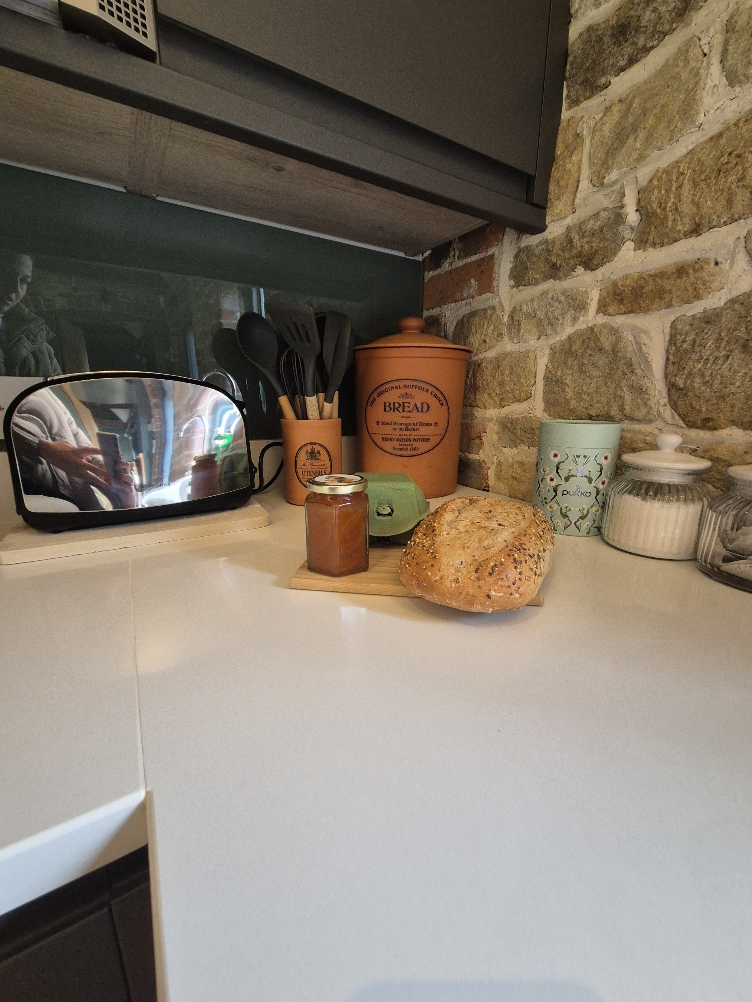 A kitchen countertop with a loaf of bread, a jar of jam, a green box, a small container of cream, and a jar of honey. Behind the items are kitchen utensils in a holder, a green box, and a container labeled 'BREAD' against a stone and brick wall.