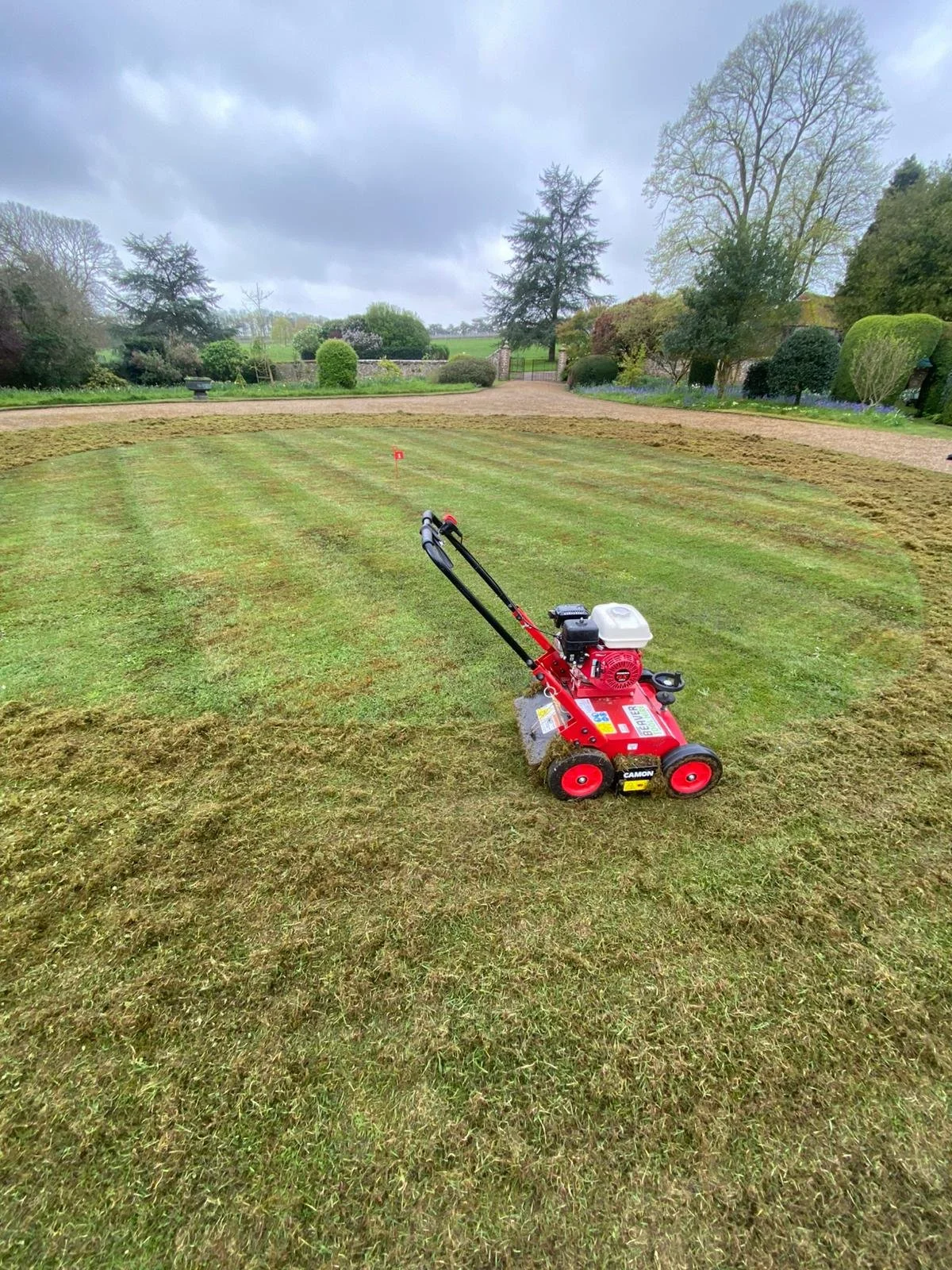 Lawn care equipment on a freshly mowed lawn in a garden with trees and bushes, cloudy sky overhead.
